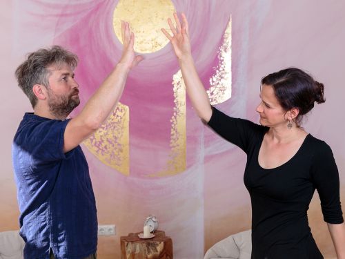 Therapeutin und Patient stehen sich mit jeweils erhobenem rechten Arm gegenüber, die Hände bewegen sich aufeinander zu, im Hintergrund bemalte Wand in Rosa- und Violetttönen