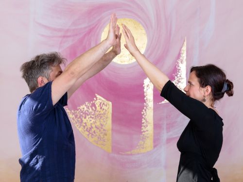 Therapeutin und Patient stehen sich mit erhobenen Armen gegenüber, die Hände berühren sich fast, im Hintergrund bemalte Wand in Rosa- und Violetttönen