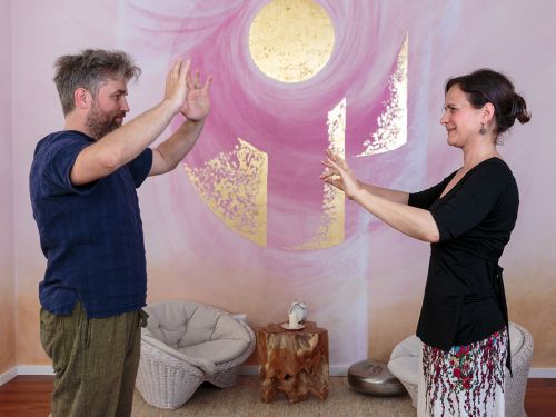Therapeutin und Patient näheren sich mit erhobenen ausgestreckten Händen an, im Hintergrund bemalte Wand in Rosa- und Violetttönen