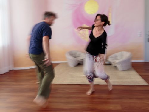 Therapeutin und Patient tanzen vor bemalter Wand in Rosa- und Violetttönen aufeinander zu, Bewegungsunschärfe