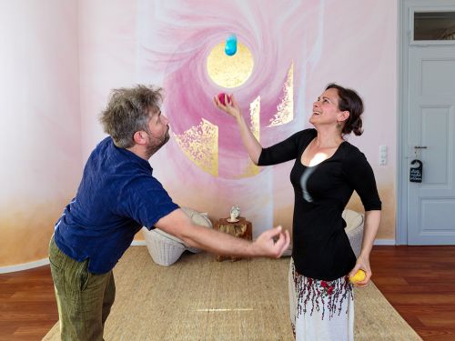 Therapeutin und Patient jonglieren gemeinsam mit bunten Filzbällen im Raum 1 vor bemalter Wand in Rosa- und Violetttönen