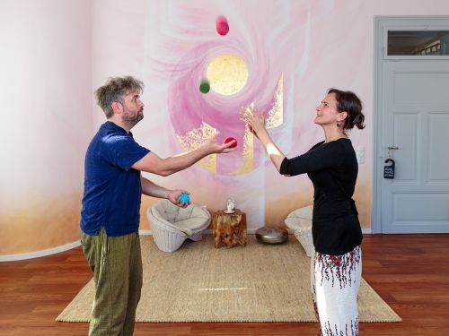 Therapeutin und Patient jonglieren gemeinsam mit bunten Filzbällen im Raum 1 vor bemalter Wand in Rosa- und Violetttönen