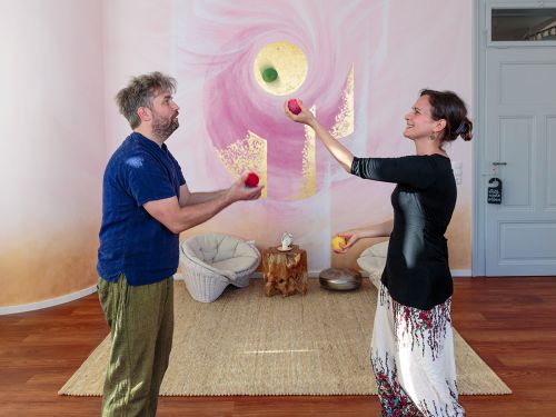Therapeutin und Patient jonglieren gemeinsam mit bunten Filzbällen im Raum 1 vor bemalter Wand in Rosa- und Violetttönen