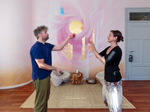 Therapeutin und Patient jonglieren gemeinsam mit bunten Filzbällen im Raum 1 vor bemalter Wand in Rosa- und Violetttönen